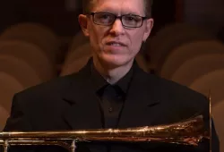 A man in dark-framed glasses and black performing clothes cradles a trombone on his lap