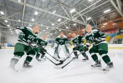 Five women in skates and green Panthers hockey gear on the ice. A goalie stands motionless in the middle. Four other players have obviously just staked into frame