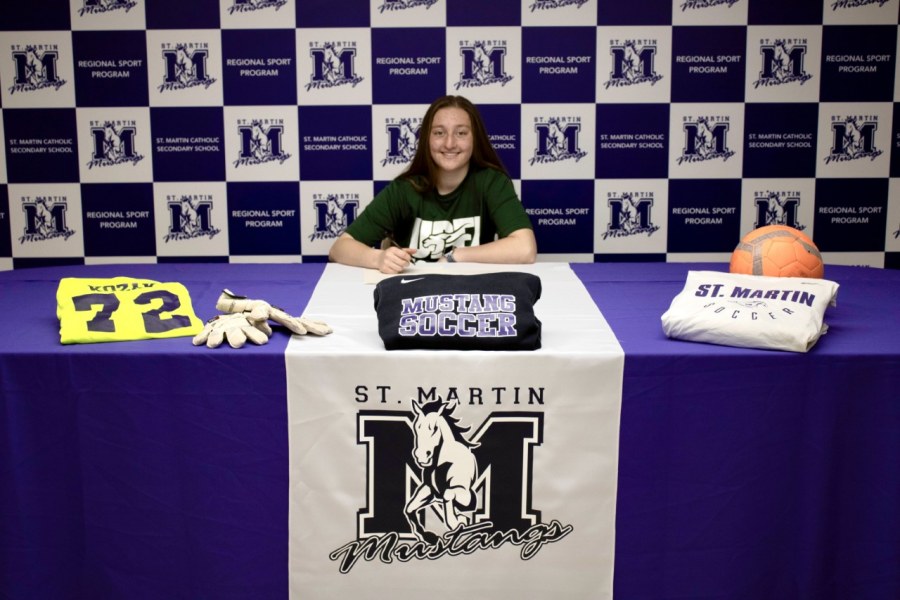 A female soccer player wearing a UPEI Panthers t-shirt sits at a table covered in logos and jerseys of her high school, St. Martins Secondary School