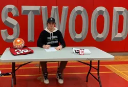 A smiling teenaged male sits at a table with the large logo fo CRESTWOOD secondary school behind him